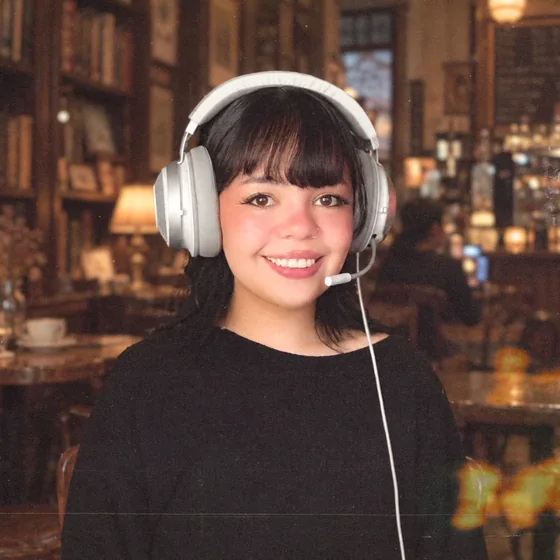 Michelle smiling in a café, wearing white headphones and a black sweater, with bookshelves in the background
